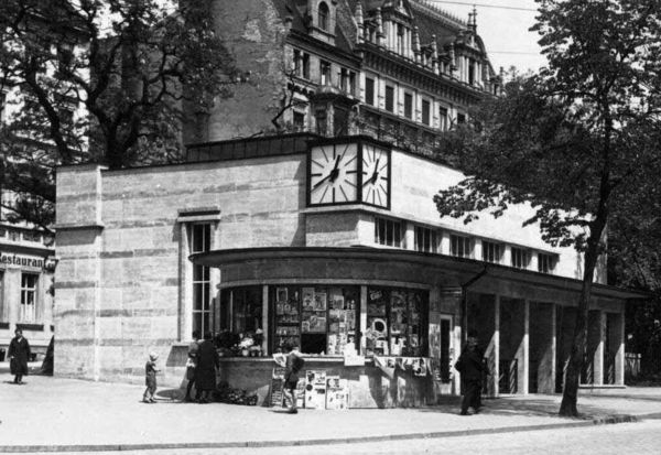 Schwarz-Weiß-Foto einer Transformatorenstation. Ein flaches Gebäude mit einer großen Uhr an der Gebäude-Ecke. An der Betrachterseite ist ein Zeitungskiosk zu sehen. Mehrere Menschen betrachten die Auslage.