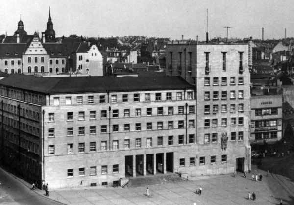 Schwarz-Weiß-Foto des Ratshofs in Halle/Saale. Ein fünfstöckiges Gebäude an einem großen Platz. An der Ecke erhebt sich ein Turm. Der Eingangsbereich ist als fünfteiliges Tor gestaltet.